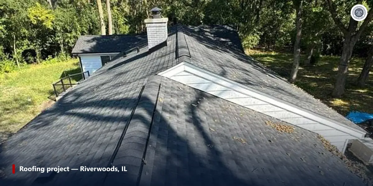 Dark gray shingle roof in Riverwoods with a long shingled ridge and a brick chimney, trees behind
