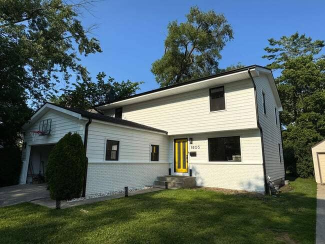 Exterior front view of a two-story residential home with white horizontal siding, white painted brick, black trim, and a vibrant yellow front door. The clean new roof features dark fascia and gutters, highlighting a complete exterior and roofing upgrade.