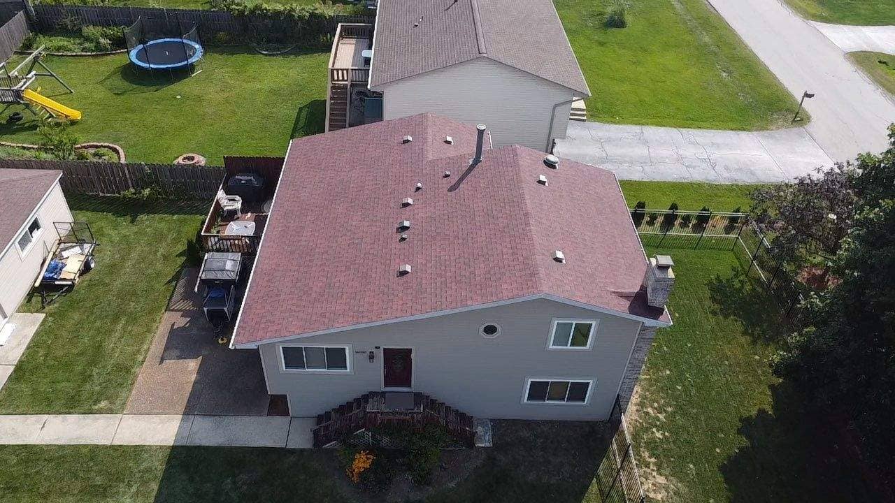 High-angle aerial drone view of a suburban house featuring a newly installed reddish-brown architectural asphalt shingle roof. The roof includes multiple exhaust vents, a plumbing stack, and a chimney on the right side. The home has light siding, a front porch entry, and is situated in a neighborhood with green lawns and neighboring houses.
