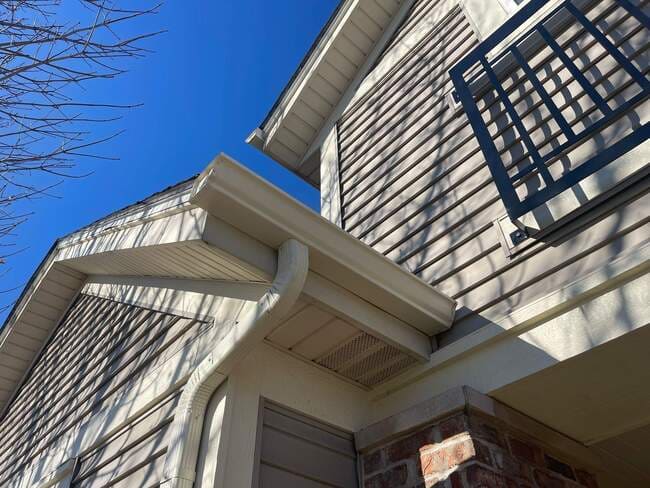 Upward-angled exterior view of a multi-story residential building featuring light siding, brick accents, a balcony railing, and a freshly installed roofline with clean white gutters and downspouts.
