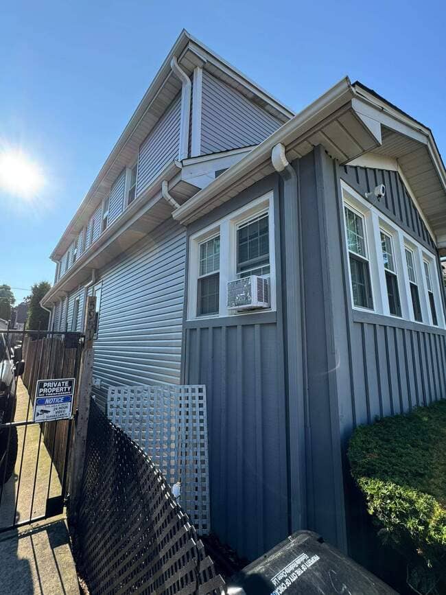 Low-angle exterior view of a multi-story residential home with light gray horizontal and vertical siding, white gutters, downspouts, and clean roof edges under a bright sunny sky.