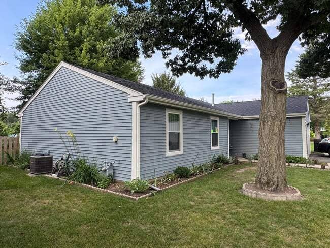 Exterior side and front view of a single-story residential home featuring light blue horizontal siding, white trim, and a newly installed dark asphalt shingle roof. A large mature tree stands in the front yard next to landscaped garden beds, highlighting a completed roofing and exterior upgrade.