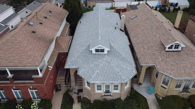 High-angle aerial view of a residential home featuring a newly installed light gray shingle roof with multiple gables and a dormer window, situated between neighboring houses.