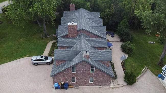 Aerial drone view looking down on a multi-gabled brick home featuring a newly installed architectural shingle roof in gray, brick chimneys, a paved driveway with a parked minivan, and surrounding green landscaping.