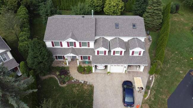 Aerial front view of a large two-story white house with red shutters, featuring a newly installed gray-brown architectural shingle roof, three front dormers, skylights, paved driveway, and lush landscaping.