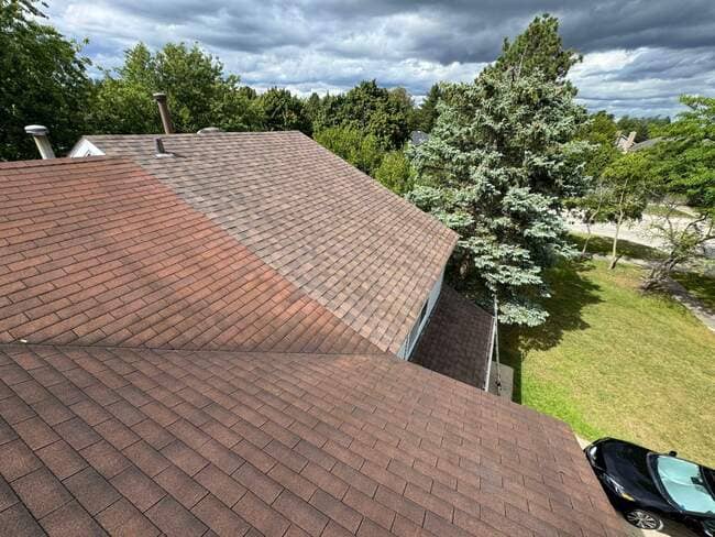 High-angle rooftop view showing multi-toned brown and reddish asphalt shingles installed across multi-sloped roof sections on a home, with evergreen trees and a car parked below against a cloudy sky.