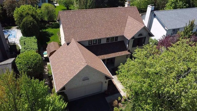 Aerial view of residential home with Owens Corning Desert Rose asphalt architectural shingles, showing full roof coverage with symmetrical ridge lines, precise edge finishing, and warm brown tones across garage and porch areas, surrounded by trees and neighboring houses