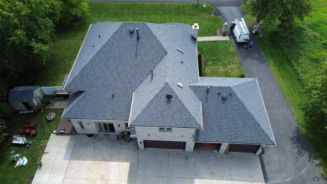 Aerial overhead view of a large residential home featuring a newly installed dark gray architectural shingle roof, multiple gables and hips, skylights, a work van in the driveway, and surrounding green lawn.