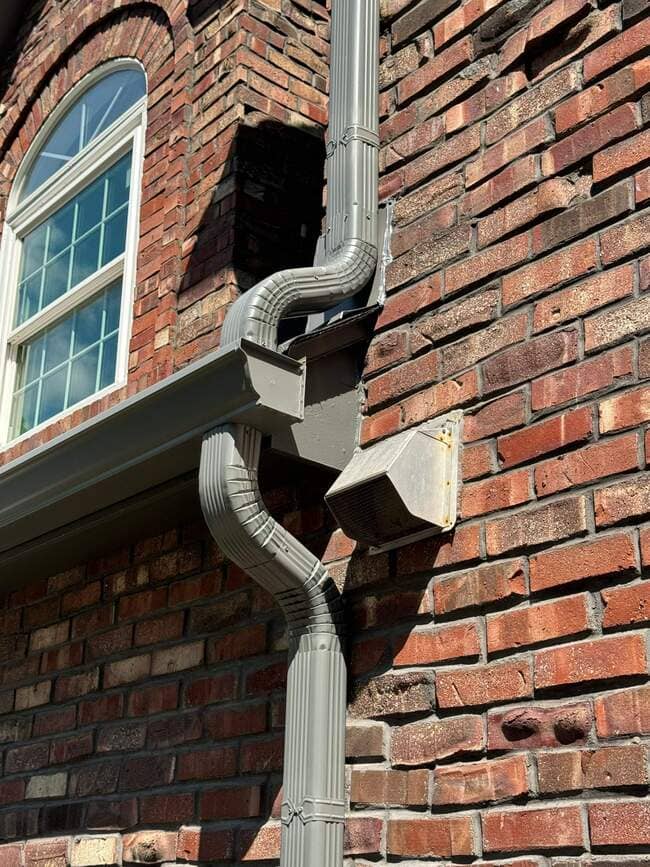 Close-up side view of a red brick house wall showing a gray aluminum downspout and gutter system installed near an arched window and exterior exhaust vent.