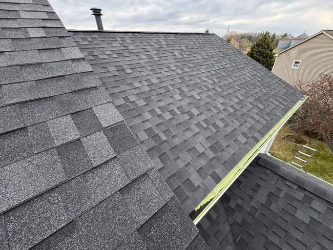 Close-up rooftop view showing high-quality dark gray architectural shingles, neat hip and ridge caps, a roof vent pipe, and green gutter line details on a residential house.