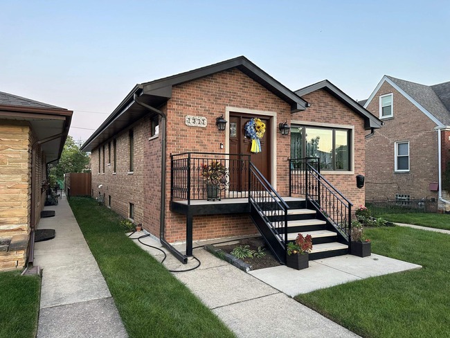 Aerial view of brick house with brown asphalt shingle roof, multi-slope design, attached garage, paved driveway, and surrounding tree.