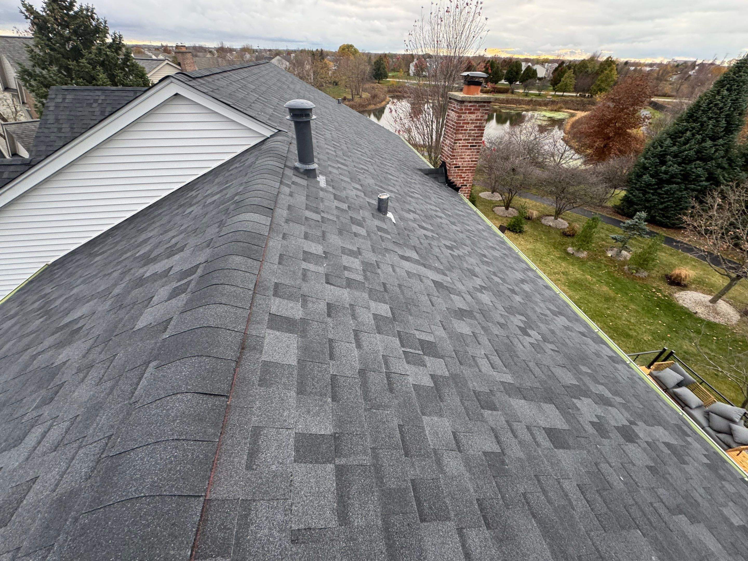 Rooftop level view of a completed residential dark gray asphalt shingle roof featuring clean lines, precise ridge caps, plumbing vent pipes, and a brick chimney. The background shows suburban scenery, trees, and a pond, showcasing professional roofing installation quality.