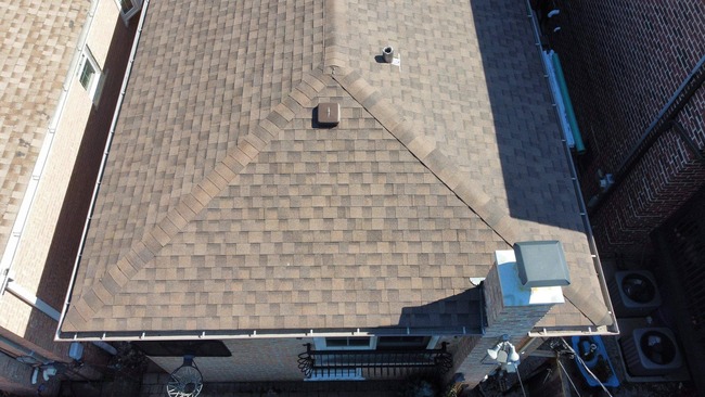Overhead drone view of a completed brown architectural asphalt shingle roof on a residential building. The hip roof features clean lines, precise ridge caps, a chimney with a cap, and integrated roof vents. The surrounding area shows adjacent brick walls and gutters along the edge of the roofing system.
