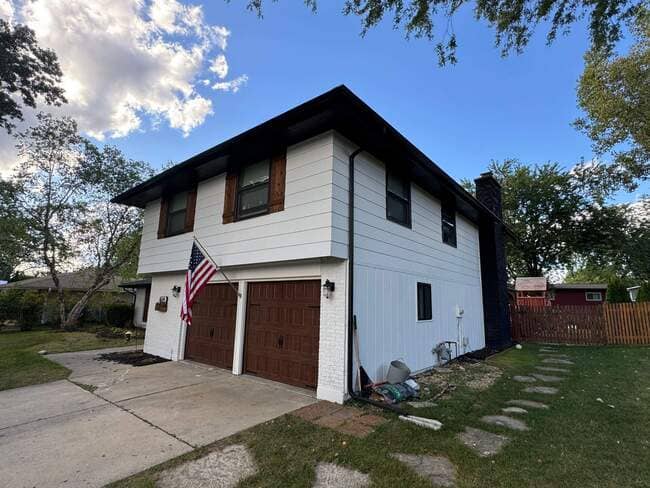 Exterior view of a two-story white siding house featuring dark brown roof eaves, a dark chimney, wooden garage doors with an American flag mounted, and a newly installed asphalt shingle roof under a blue sky with soft clouds.