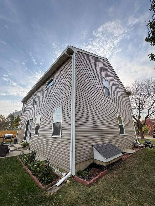 Low-angle upward exterior view of a two-story residential house with beige vinyl siding, white gutters and downspouts, roof overhangs, and a shingled basement entryway hatch against a bright sky.