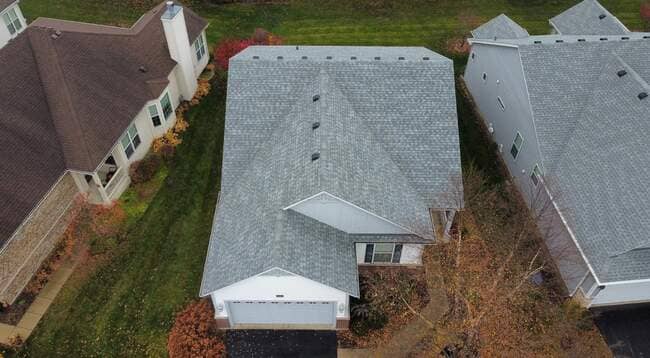 High-angle overhead drone view of a suburban house featuring a newly installed light gray architectural shingle roof, roof ridge vents, attached garage, and surrounding green lawns.