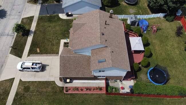 Aerial top-down drone view of a multi-level suburban house featuring a newly installed tan architectural shingle roof, skylight, chimney, work truck parked in driveway, back deck, gazebo, trampoline, and lawn.