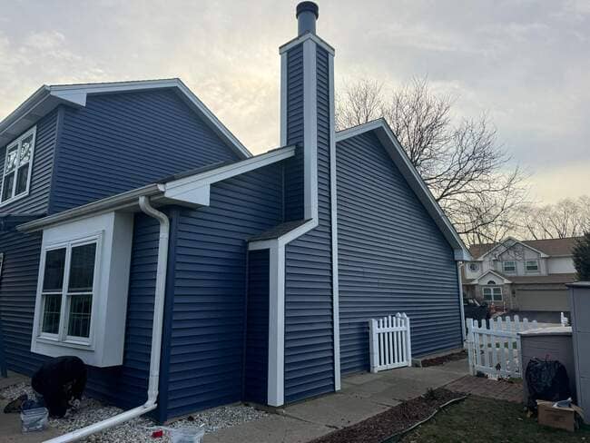 Aerial drone view of a completed dark gray asphalt shingle roof installation on a single-story residential home nestled within dense green trees. The clean, modern roof system features vent stacks and a central chimney, showcasing a high-quality, professional roof replacement. Three service vehicles are visible in the gravel driveway next to the house.