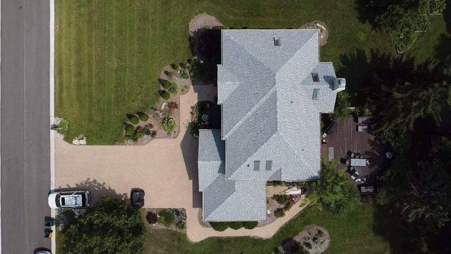 Aerial view of residential property with light-colored asphalt shingle roof featuring multiple sections, uniform shingle layout, clean ridge lines, and full coverage over garage and main structure, surrounded by landscaped lawn, driveway, and adjacent road