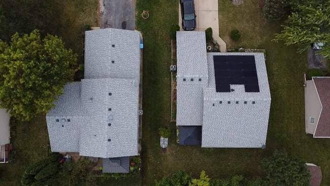 Top-down aerial drone view of two adjacent residential houses featuring newly installed light gray asphalt shingle roofs, with one of the properties showcasing a clean integration of rectangular black solar panels.