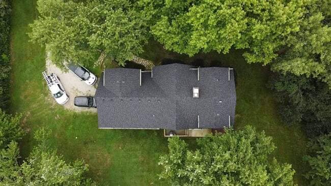 Aerial drone view of a completed dark gray asphalt shingle roof installation on a single-story residential home nestled within dense green trees. The clean, modern roof system features two vent stacks and a prominent white chimney, showcasing a high-quality, professional roof replacement. Three service vehicles are visible in the gravel driveway next to the house.