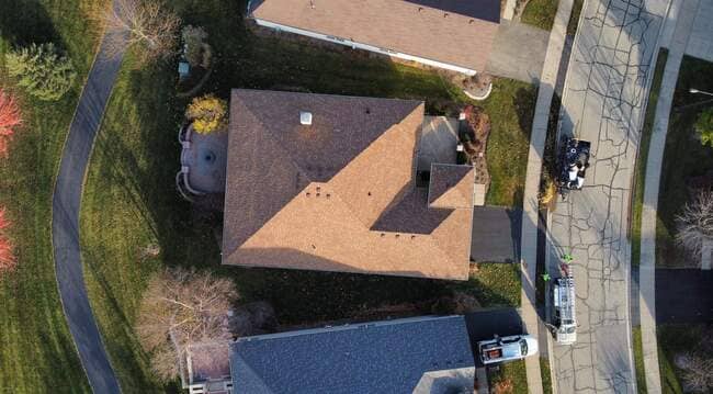Top-down aerial drone view of a suburban house featuring a newly installed brown architectural asphalt shingle roof. The multi-section hip roof displays clean lines, ventilation pipes, and a small upper section. A contractor's work vehicle and equipment are parked on the street alongside the property, highlighting an active roof replacement project.