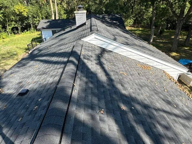 Elevated rooftop view of a single-family residential house featuring dark gray asphalt architectural shingles, a clean ridge vent, a brick chimney, and white trim under shade trees.