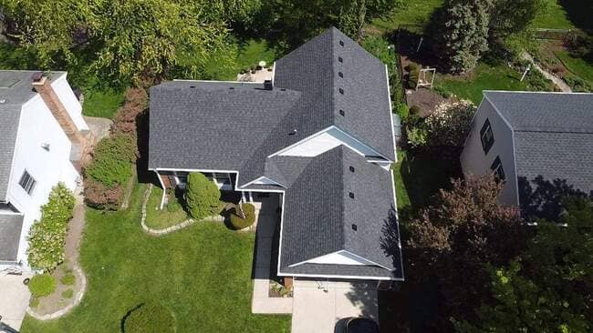 Aerial top-down drone view of a suburban house featuring a newly installed dark gray architectural shingle roof, roof vents, lush green yard, concrete walkway, and surrounding trees.