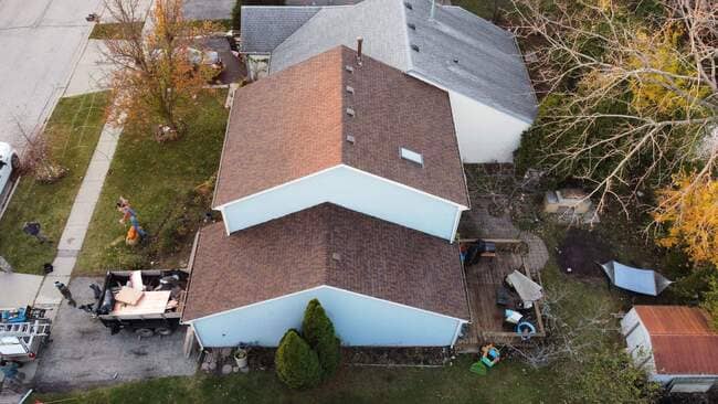Aerial drone view of a multi-level suburban home featuring a newly installed brown architectural asphalt shingle roof. The roof includes a skylight, plumbing vent stacks, and clean ridge lines. A contractor's trailer and equipment are visible in the driveway, indicating an active or recently finished roof replacement project.