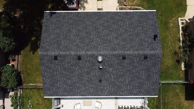 Aerial view of residential home with dark gray asphalt shingle roof, multiple roof vents, a skylight, green lawn, and paved patio with outdoor furniture.