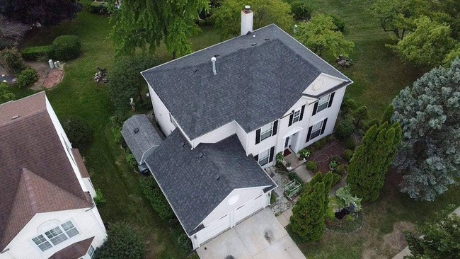 Aerial shot of a two-story white residential house with a newly installed dark gray shingle roof. The image captures the house and surrounding green landscape, a two-car garage, and a driveway with black decorative shutters on the windows.