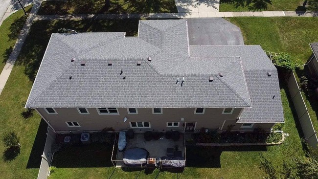 Aerial view of a large suburban L-shaped house with a newly installed light grey asphalt shingle roof. The image captures the entire roof and surrounding property, including a patio with outdoor furniture, green lawns, and a driveway.