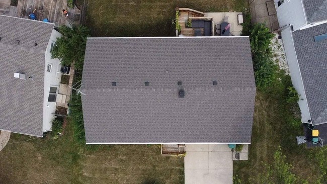 Aerial view of a suburban house with a newly installed dark gray asphalt shingle roof. The image is a top-down shot showing the rectangular shape of the roof, the surrounding green lawn, a driveway, and adjacent houses with decks and patios.