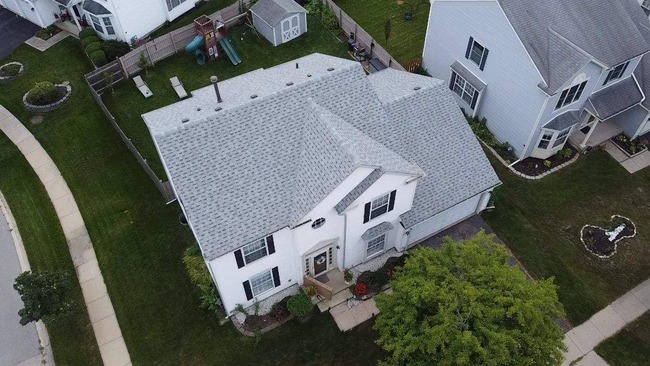 Aerial view of a white, two-story suburban house with a newly installed light grey asphalt shingle roof. The image shows the entire home, including a front yard with a walkway, a driveway, a backyard with a playground set and shed, and adjacent neighboring houses.