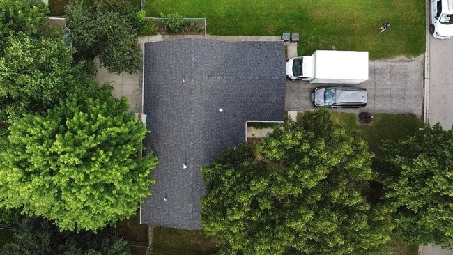 Aerial view of a newly installed, dark charcoal gray asphalt shingle roof on a single-story residential home. The professional installation is clean and uniform, surrounded by lush green lawns and large trees, with a driveway containing a white box truck and a black SUV.