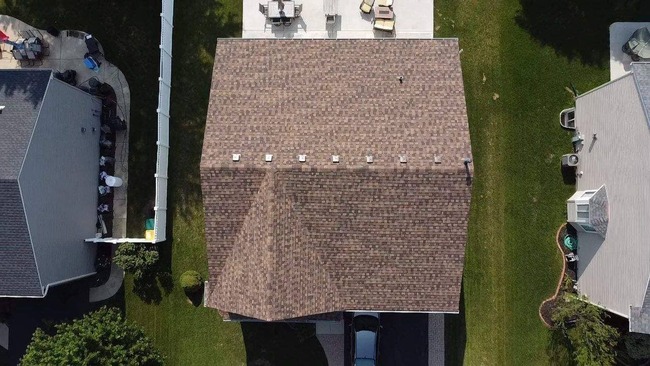 Aerial view of a newly installed residential roof featuring brown shingles on a suburban home. The image shows the clean, uniform installation of the shingles, the vent pipes, and the well-maintained lawn and backyard patio area surrounding the house.
