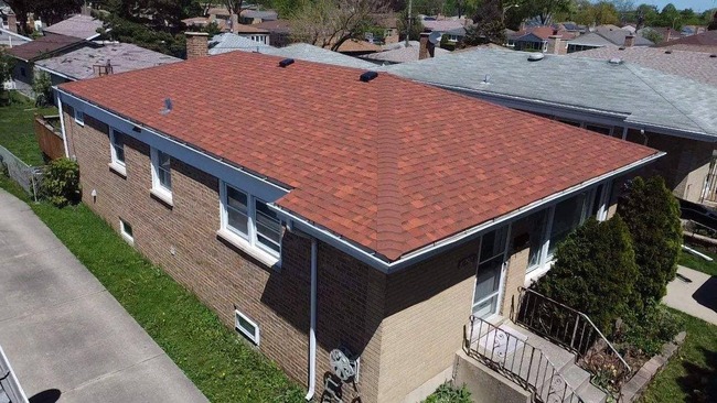 Aerial view of a newly installed reddish-brown asphalt shingle roof on a brick and tan-sided single-story suburban home. The image captures the entire roof, showcasing the uniform installation, as well as the surrounding green lawn, driveway, and neighboring houses under a clear sky.