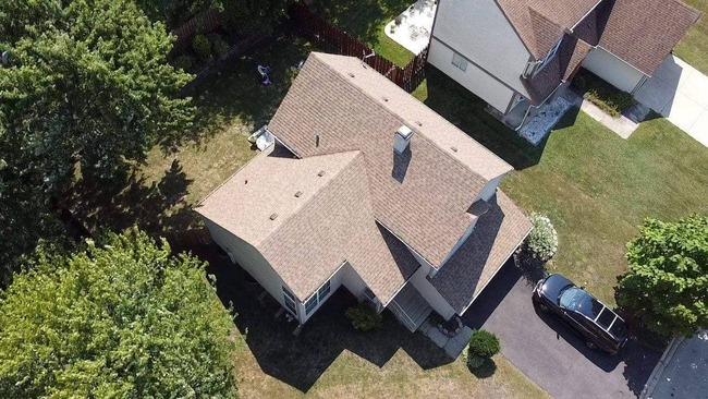 Aerial view of a newly installed brown asphalt shingle roof overlay on a two-story beige suburban house. The image captures the home's multi-tonal roof, surrounding green lawn, a driveway with a black car, and neighboring houses, highlighting the quick and professional upgrade.