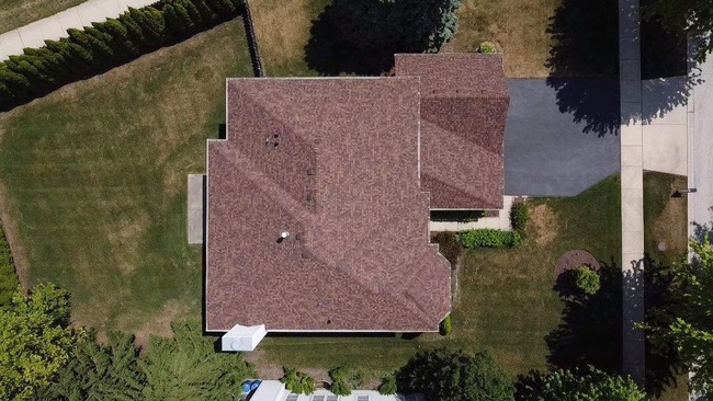 Aerial view of residential home with brown asphalt shingle roof featuring multi-angle design, two roof vents, attached garage, paved driveway, landscaped lawn, and surrounding trees - professionally installed for durability and visual appeal