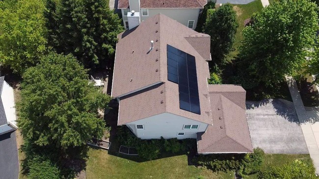 Aerial view of residential home with uniquely colored brown asphalt shingle roof, solar panels installed in rectangular formation, surrounding trees, visible driveway, and neighboring houses
