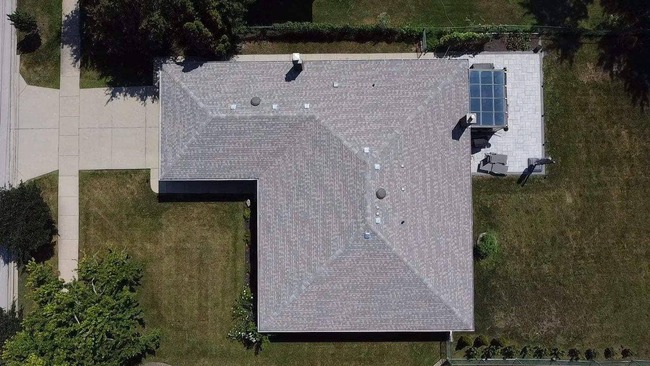 Aerial view of a suburban L-shaped house with a newly installed light grey asphalt shingle roof. The image captures the entire roof and surrounding property, including a patio with outdoor furniture, green lawns, and a driveway.