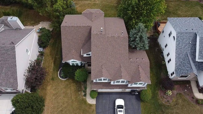 Aerial view of a large two-story suburban house with a newly installed multi-tonal brown asphalt shingle roof. The image captures the entire roof and surrounding landscape, including green lawns, neighboring homes, and a driveway with a white car.
