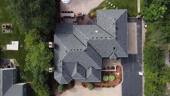 Aerial view of a large residential house with a complex roof structure, featuring a new dark gray asphalt shingle roof resulting from a fully-covered insurance claim. The image captures the property, including a paver driveway, a back patio, and surrounding landscaping.